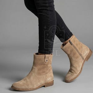 May include: Tan suede ankle boots with perforated detailing and a side zipper. The boots have a low heel and a rounded toe. The boots are worn with black jeans. The boots are a neutral color and can be worn with many outfits.