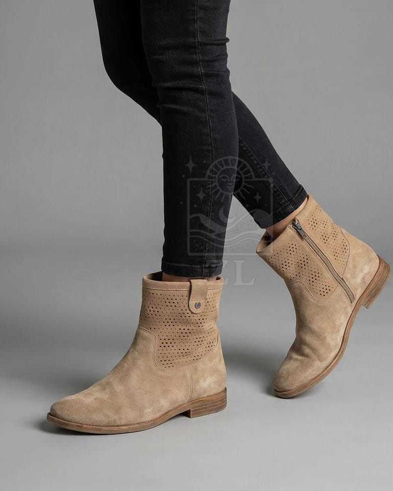 May include: Tan suede ankle boots with perforated detailing and a side zipper. The boots have a low heel and a rounded toe. The boots are worn with black jeans. The boots are a neutral color and can be worn with many outfits.