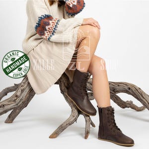 May include: Brown leather lace-up boots with a side zipper. The boots have a flat sole and are worn by a person sitting on a wooden branch. The boots are part of the Zalle Shop collection, as indicated by the logo in the image.
