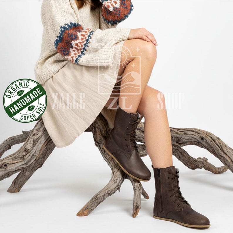 May include: Brown leather lace-up boots with a side zipper. The boots have a flat sole and are worn by a person sitting on a wooden branch. The boots are part of the Zalle Shop collection, as indicated by the logo in the image.