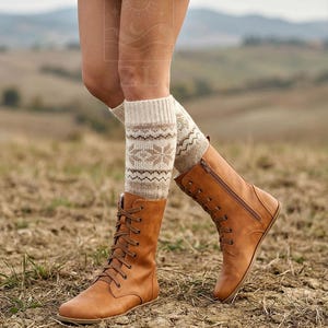 May include: A pair of tan leather lace-up boots with a side zipper, paired with patterned knit leg warmers. The boots have a flat sole and are worn outdoors on a dirt surface. The leg warmers are beige with a geometric design.