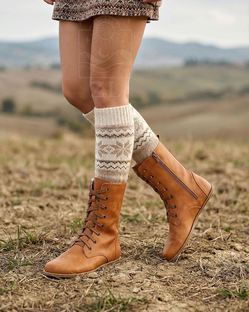 May include: A pair of tan leather lace-up boots with a side zipper, paired with patterned knit leg warmers. The boots have a flat sole and are worn outdoors on a dirt surface. The leg warmers are beige with a geometric design.