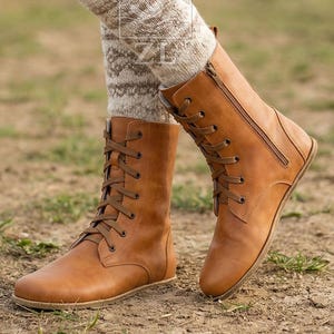 May include: A pair of tan leather boots with laces and a side zipper. The boots are mid-calf height and have a flat sole. The boots are worn with patterned socks. The boots are on a dirt surface.