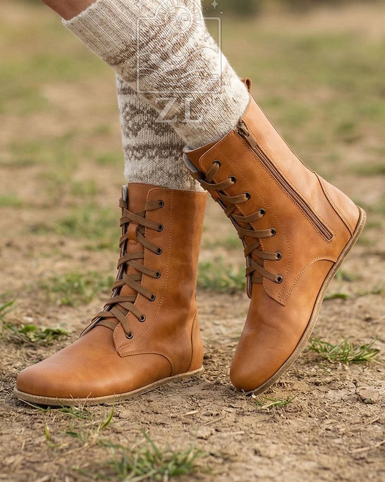 May include: A pair of tan leather boots with laces and a side zipper. The boots are mid-calf height and have a flat sole. The boots are worn with patterned socks. The boots are on a dirt surface.