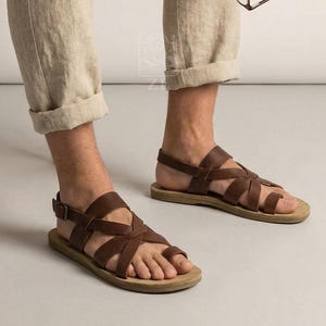 Puede incluir: Sandalias de cuero marrón con múltiples correas y cierre de hebilla. Las sandalias tienen una suela marrón claro y se usan con pantalones de lino beige. La imagen muestra las sandalias en los pies.