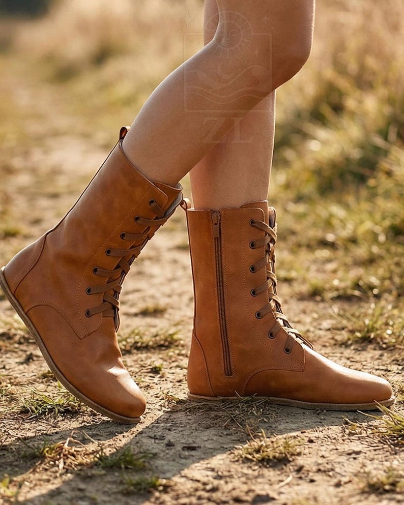 May include: Tan leather boots with a lace-up front and side zipper. The boots have a flat sole and are mid-calf height. The boots are worn outdoors on a dirt path.