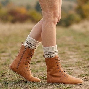May include: A person wearing tan leather lace-up boots with a side zipper and patterned socks. The boots have a flat sole and are worn with beige and brown patterned socks. The background is a grassy field with trees.