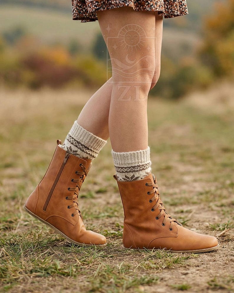 May include: A person wearing tan leather lace-up boots with a side zipper and patterned socks. The boots have a flat sole and are worn with beige and brown patterned socks. The background is a grassy field with trees.
