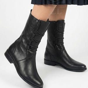 Puede incluir: Botas de cuero negro de media caña con tacón bajo. Las botas tienen un diseño con cordones en la parte superior de la caña y una puntera redondeada. Las botas se muestran desde las rodillas hacia abajo.
