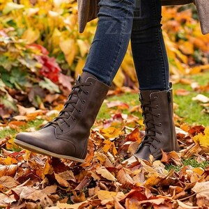 May include: Brown leather lace-up boots with a side zipper. The boots are worn with dark blue jeans and are surrounded by fallen autumn leaves in shades of yellow, orange, and red. The boots have a flat sole.