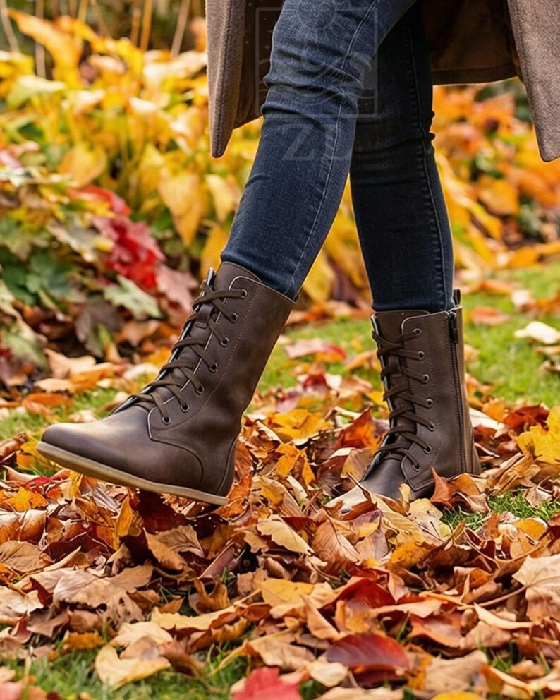 May include: Brown leather lace-up boots with a side zipper. The boots are worn with dark blue jeans and are surrounded by fallen autumn leaves in shades of yellow, orange, and red. The boots have a flat sole.
