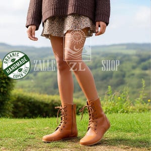 May include: A pair of light brown leather lace-up boots with a side zipper. The boots have a round toe and a flat sole. The boots are worn on a grassy field. The boots are labeled "Organic Handmade Coper Sole".