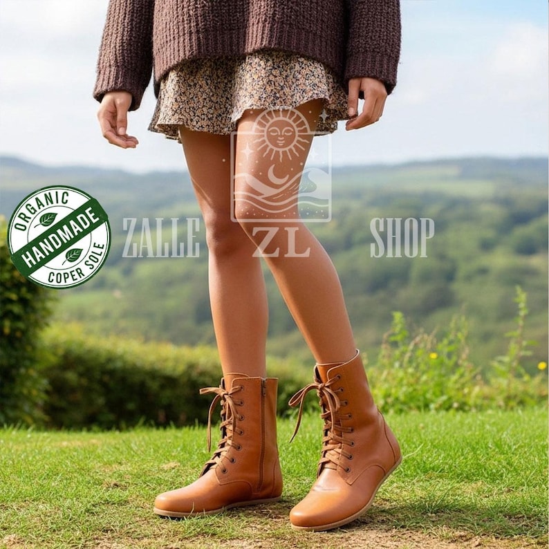 May include: A pair of light brown leather lace-up boots with a side zipper. The boots have a round toe and a flat sole. The boots are worn on a grassy field. The boots are labeled "Organic Handmade Coper Sole".