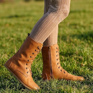 May include: Tan-colored lace-up boots with a side zipper. The boots are made of a smooth material and have a flat sole. The boots are worn with patterned tights, and the background is a grassy field.