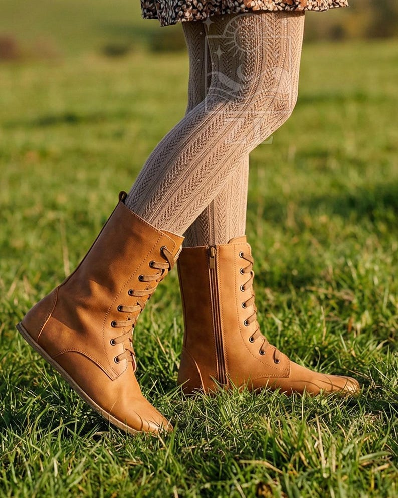 May include: Tan-colored lace-up boots with a side zipper. The boots are made of a smooth material and have a flat sole. The boots are worn with patterned tights, and the background is a grassy field.