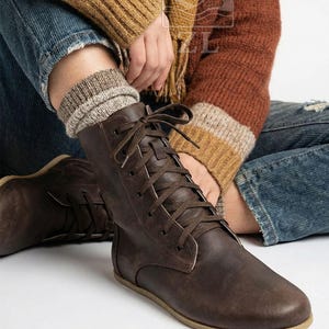 May include: Brown leather lace-up boots with a flat sole. The boots have a worn, textured appearance and are laced up with brown laces. The boots are paired with a pair of gray and beige socks. The boots are a casual style.