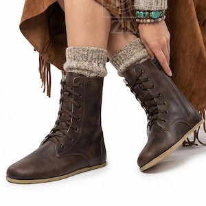 May include: Brown leather lace-up boots with a side zipper and tan soles. The boots are worn with beige knit leg warmers. The image shows a close-up of the boots and part of the person's legs.