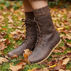 May include: Brown leather lace-up boots with light-colored soles. The boots are worn with brown socks and are surrounded by fallen autumn leaves on the ground. The boots have a classic, timeless design.