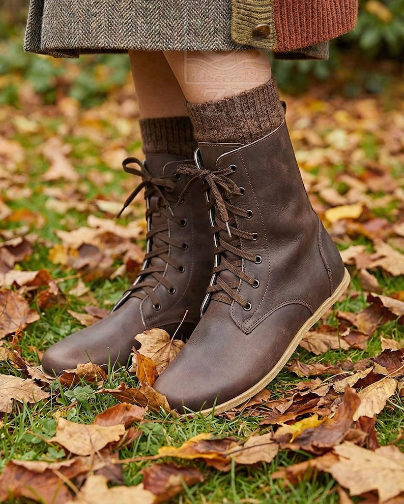 May include: Brown leather lace-up boots with light-colored soles. The boots are worn with brown socks and are surrounded by fallen autumn leaves on the ground. The boots have a classic, timeless design.