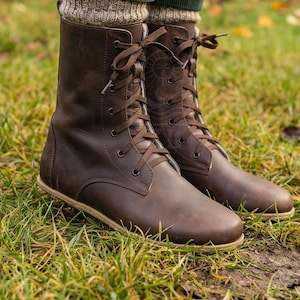 May include: Brown leather lace-up boots with a round toe and a flat sole. The boots have a mid-calf height and are laced up with brown laces. The boots are worn with green corduroy pants and beige knit socks. The boots are on a grassy surface.