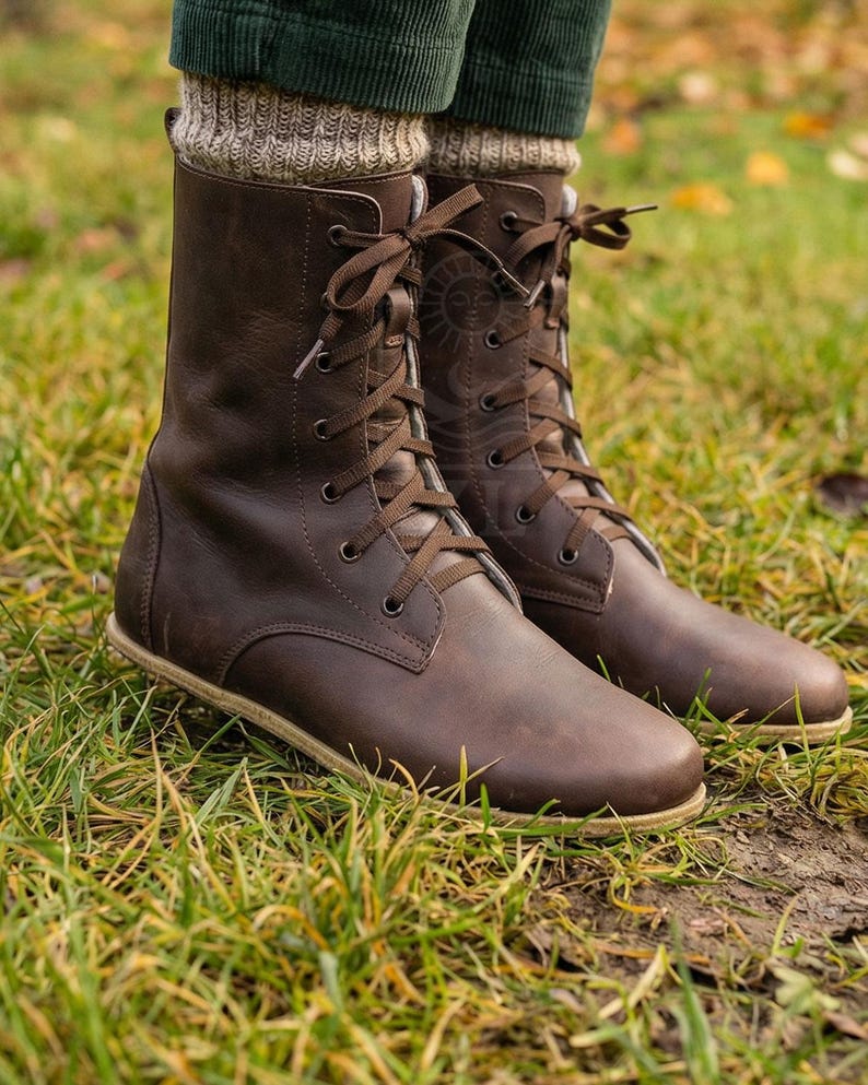 May include: Brown leather lace-up boots with a round toe and a flat sole. The boots have a mid-calf height and are laced up with brown laces. The boots are worn with green corduroy pants and beige knit socks. The boots are on a grassy surface.