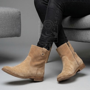 May include: Tan suede ankle boots with perforated detailing. The boots have a side zipper and a small buckle detail. The boots are worn with black jeans.