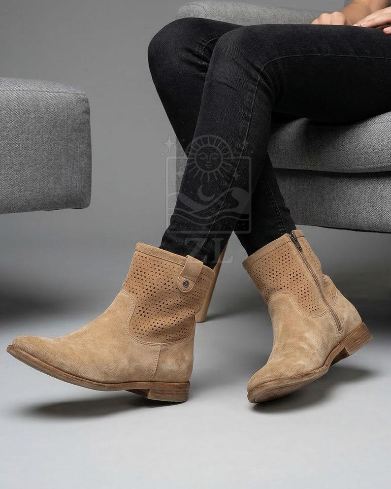 May include: Tan suede ankle boots with perforated detailing. The boots have a side zipper and a small buckle detail. The boots are worn with black jeans.