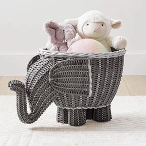 Puede incluir: Una cesta de almacenamiento gris en forma de elefante de mimbre llena de peluches. La cesta tiene un diseño tejido y está sostenida por cuatro patas pequeñas. En el interior hay un elefante gris, un cordero blanco y una bola de felpa rosa.