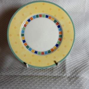 May include: A round ceramic plate with a yellow background and a white centre. The plate features a border of colourful squares and small orange dots. The rim is a light green colour.