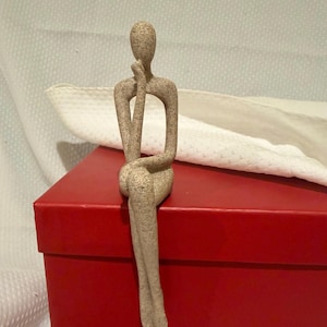 May include: A beige, speckled, abstract human figurine in a seated, contemplative pose. The figure has an elongated, smooth form with a rounded head, resting its chin on one hand while the other arm rests on its crossed legs, creating a hollow space.