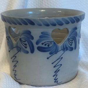 May include: A cylindrical stoneware crock featuring a light grey base with hand-painted blue floral motifs and heart-shaped cutouts along the upper rim. The rim is decorated with a rope-like texture, and the body displays blue squiggly line accents.
