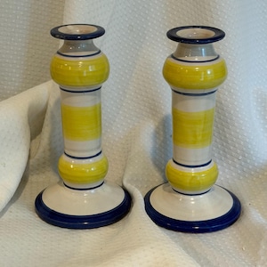 May include: A pair of ceramic candle holders featuring a cream-colored base with horizontal yellow bands and thin dark blue rings. Each holder has a flared circular base, a central cylindrical stem with two rounded sections, and a wide rim.