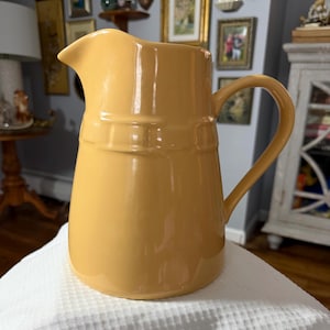 May include: A golden-yellow ceramic pitcher with a curved handle and a spout. The pitcher has a smooth, glossy finish and a decorative band around the middle. It is a classic kitchenware item.