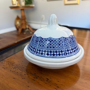 May include: A white ceramic butter dish with a blue geometric pattern on the lid. The dish has a rounded base and a domed lid with a small handle. The pattern is a repeating design of squares and diamonds.