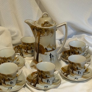 May include: A porcelain coffee set featuring a tall lidded coffee pot and four matching cups with saucers. Each piece is decorated with a brown and white landscape scene depicting trees, small buildings, and figures walking in a snowy setting.
