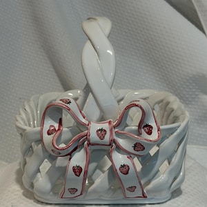 Peut inclure: Un panier en céramique blanche avec un design en treillis tressé et une anse torsadée unique. L'avant présente un nœud décoratif en céramique avec un bord rose et un motif répétitif de petites fraises rouges peintes sur toute la surface.