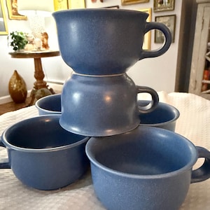 以下が含まれることがあります： 取っ手付きの青いセラミックマグカップ5個。マグカップは積み重ねて配置されています。マグカップは斑点のある質感と丸みを帯びた形状をしています。マグカップはくすんだ青色です。マグカップはさまざまな位置にあり、積み重ねられたものと直立したものがあります。