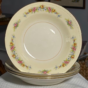 May include: A stack of cream-colored soup bowls with a floral design. The bowls have a gold rim and a decorative border of pink, yellow, and blue flowers. The bowls are stacked on top of each other.