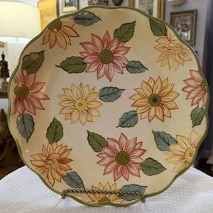 May include: A decorative ceramic plate with a scalloped edge, featuring a cream-colored background adorned with painted sunflowers in shades of yellow, pink, and orange. Green leaves and a green border complete the design. The plate is displayed on a metal stand.