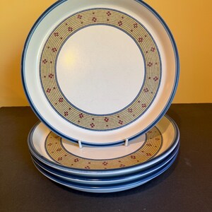 May include: A stack of four white ceramic plates with a blue rim and a decorative band of tan and red geometric patterns. The plates are stacked on a black surface against a yellow background.