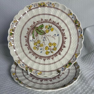 May include: Two white ceramic plates with a scalloped edge. Each plate features a floral design with yellow flowers, green leaves, and a brown border. The plates have a woven basket-like texture.