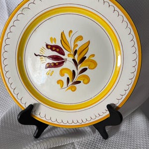 Könnte beinhalten: Ein weißer Keramikteller mit gewelltem Rand und einem gelb-braunen Blumendesign. Der Teller hat einen gelben Rand und wird auf einem schwarzen Ständer präsentiert. Das florale Design zeigt gelbe und kastanienbraune Blätter.