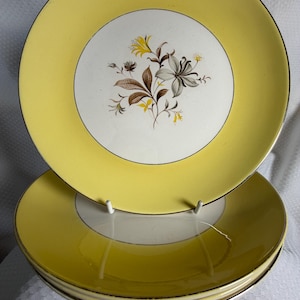 May include: A stack of yellow and white ceramic plates with a floral design. The plates have a yellow border and a white centre with a brown, grey, and yellow floral pattern. The plates are stacked on a white surface.