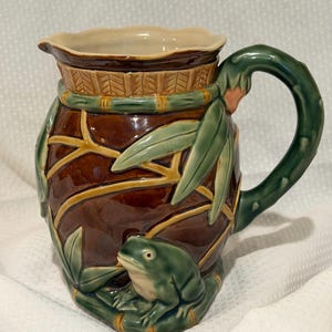 May include: A ceramic pitcher featuring a brown body with a raised green frog sitting on a bamboo-style base. The pitcher has a green bamboo-shaped handle, green leaves, and a textured rim. The interior is cream-colored with a scalloped edge.