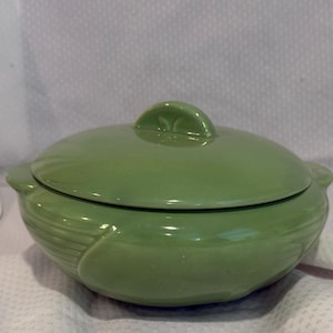 May include: A sage green ceramic casserole dish with a matching lid. The round vessel features a smooth, glossy finish and integrated side handles. The lid includes a central arched handle, and the exterior base displays subtle vertical ribbing.