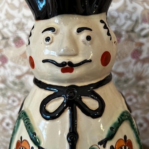 Puede incluir: Figura de chef de cerámica con sombrero negro y rostro pintado. La figura tiene una pajarita negra y un patrón floral decorativo con detalles en naranja, rojo y verde. Es un objeto decorativo.