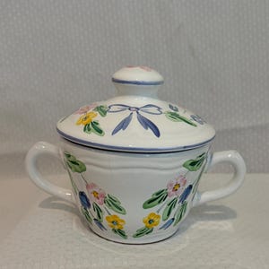 May include: A white ceramic sugar bowl with two handles and a matching lid. The exterior features a hand-painted floral pattern with pink, yellow, and blue flowers, green leaves, and a blue ribbon motif. The lid has a small knob handle on top.