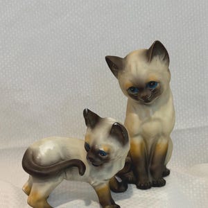 Puede incluir: Dos figuras de gatos siameses con cuerpos de color crema y orejas, caras, colas y patas de color marrón oscuro. Un gato está de pie de perfil mirando hacia atrás, mientras que el otro gato está sentado erguido y mirando al frente con ojos azules.
