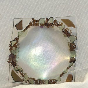 May include: A square glass dish featuring a central iridescent, pearlescent circular area. The border is decorated with a mosaic of small, irregular brown and light green glass fragments, with larger brown glass pieces at the four corners.