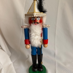 May include: A wooden nutcracker figure wearing a white triangular hat with gold trim and a black feather. The figure has a white faux fur beard, blue jacket with red cuffs, blue trousers, and black boots, standing on a round green base.